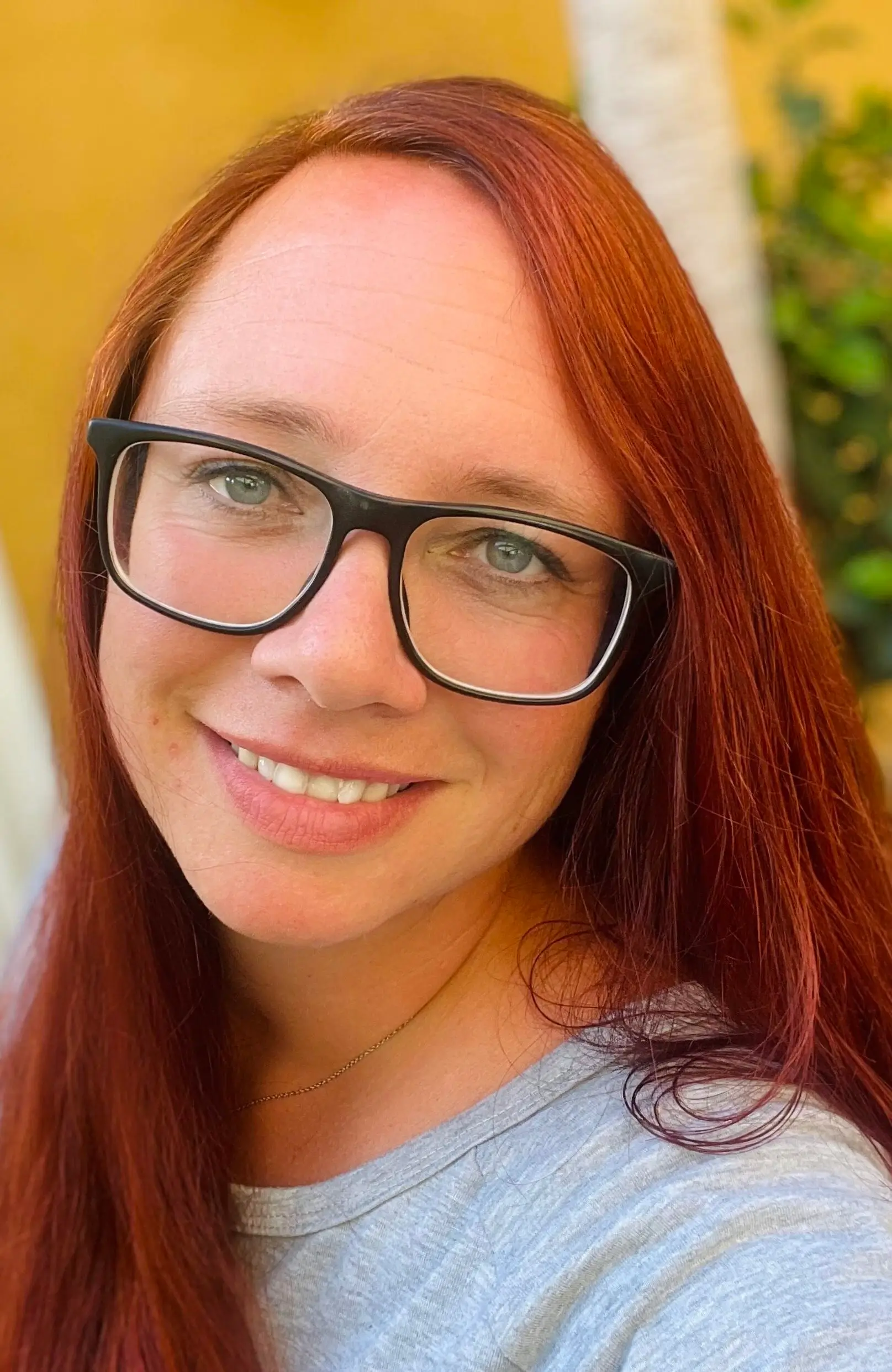 Mujer sonriente con cabello largo y rojo, usando gafas, en un fondo natural. La imagen refleja una expresión amigable y positiva.
