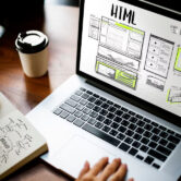 Diseño de interfaz de usuario en pantalla de laptop mostrando esbozos de HTML, con una persona tomando notas y un café al lado.