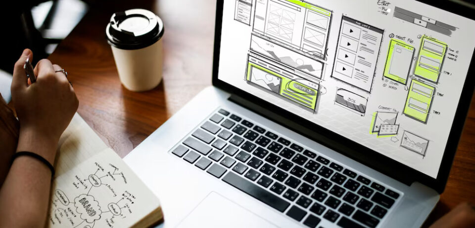 Diseño de interfaz de usuario en pantalla de laptop mostrando esbozos de HTML, con una persona tomando notas y un café al lado.