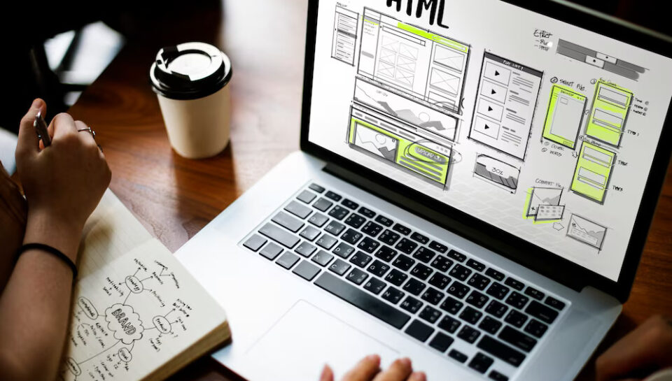 Diseño de interfaz de usuario en pantalla de laptop mostrando esbozos de HTML, con una persona tomando notas y un café al lado.